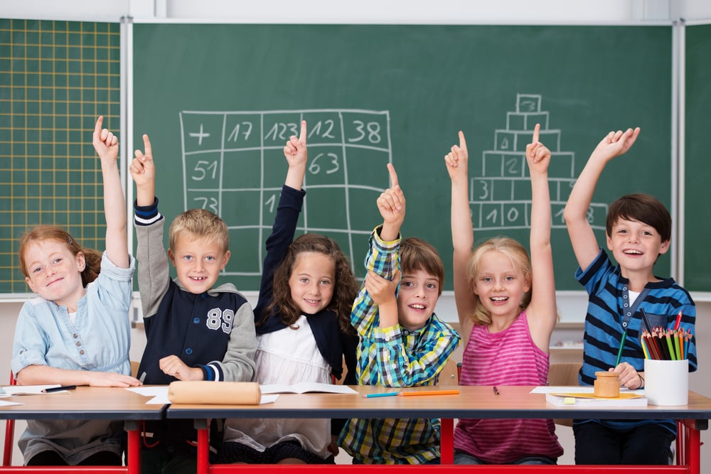 Shields Childcare Supplies Image of young kids in class sitting in a row at their desk raising their hands in the air to show the know the answer to a question